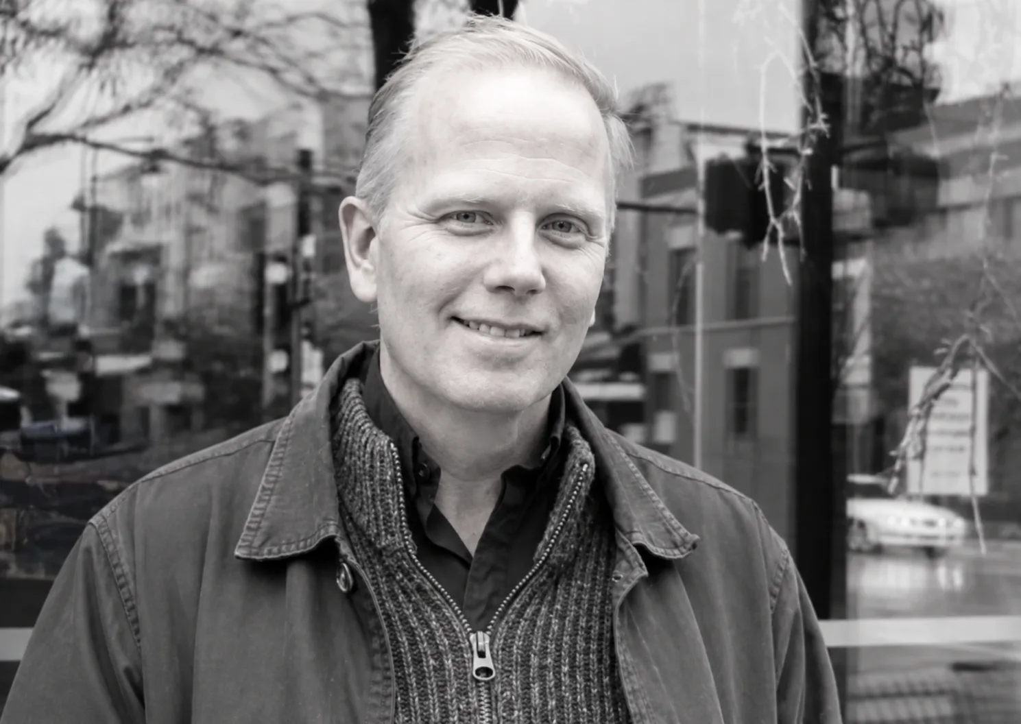 Black and white portrait of Jamie Parsley in a jacket and ribbed sweater in front of a shop window reflecting a city street