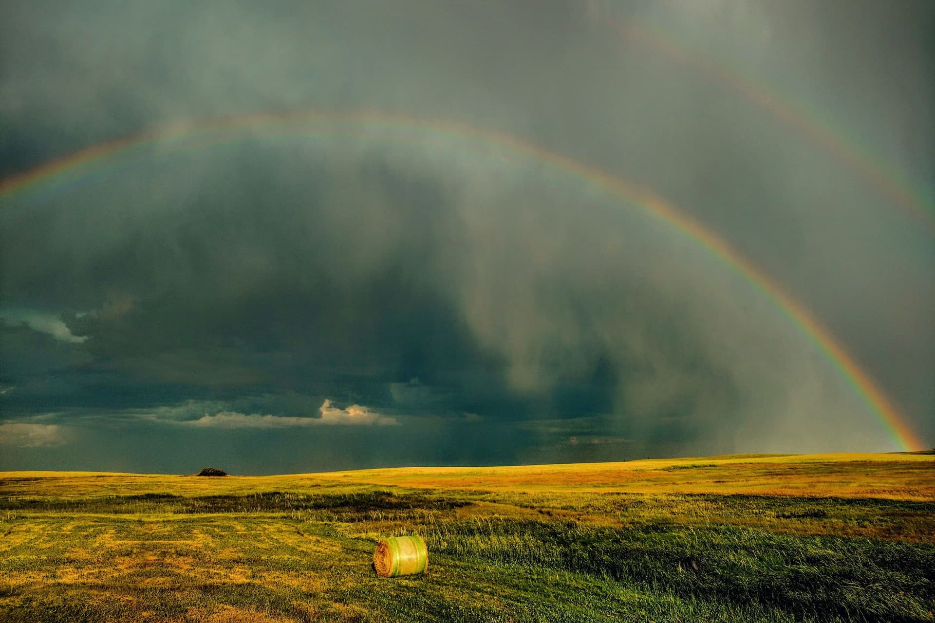 A double rainbow over a hayfield with a single round bale under a dark storm sky
