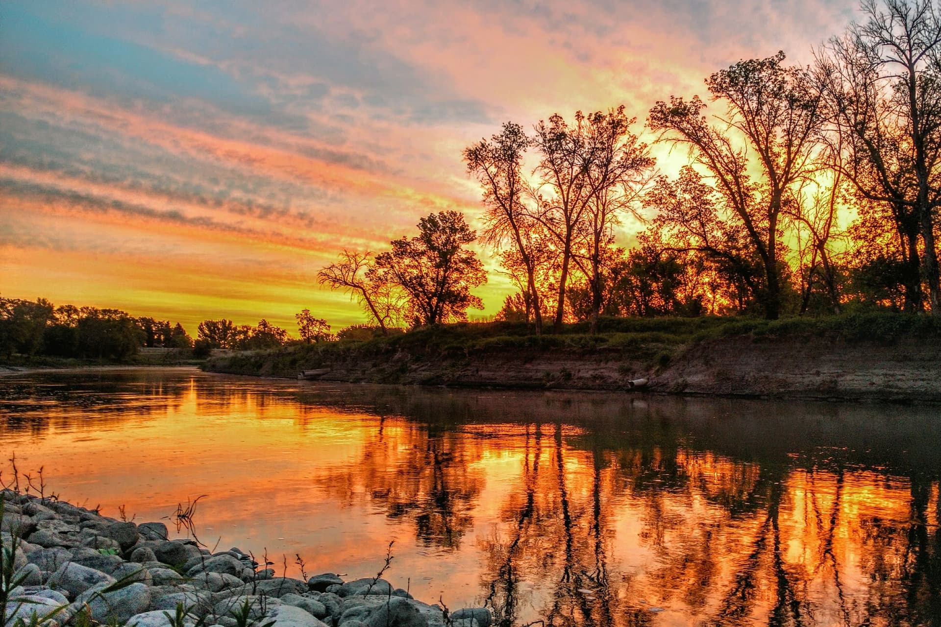 Trees along a riverbank silhouetted against an orange and pink sunset, mirrored in still water